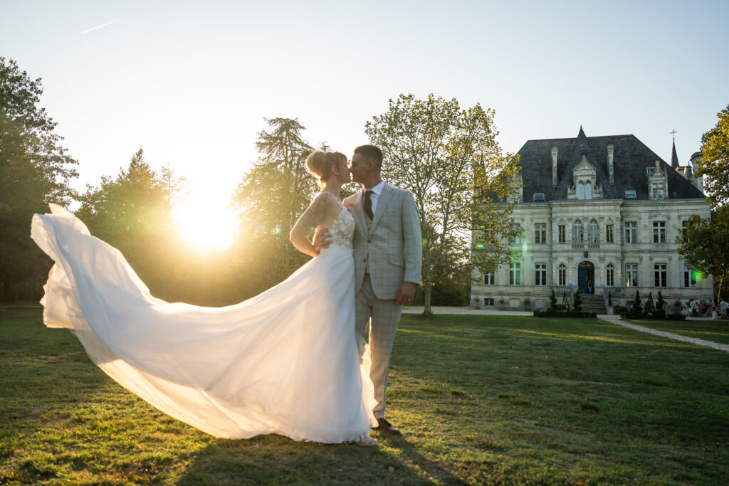 Wedding couple kissing in front of a grand house at sunset with the bride’s dress flowing in the light.