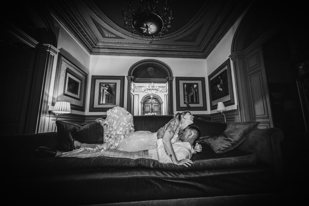Black and white photo of a wedding couple lying on a sofa, embracing and laughing in an ornate historic room.