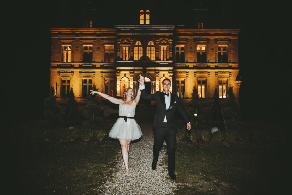 Wedding couple walking hand in hand happily in front of a large historic building lit with warm lights.