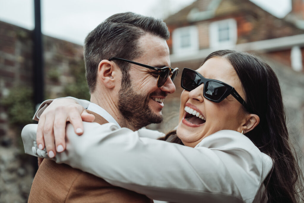 Wedding couple wearing suits and sunglasses hugging as they laugh together outdoors.
