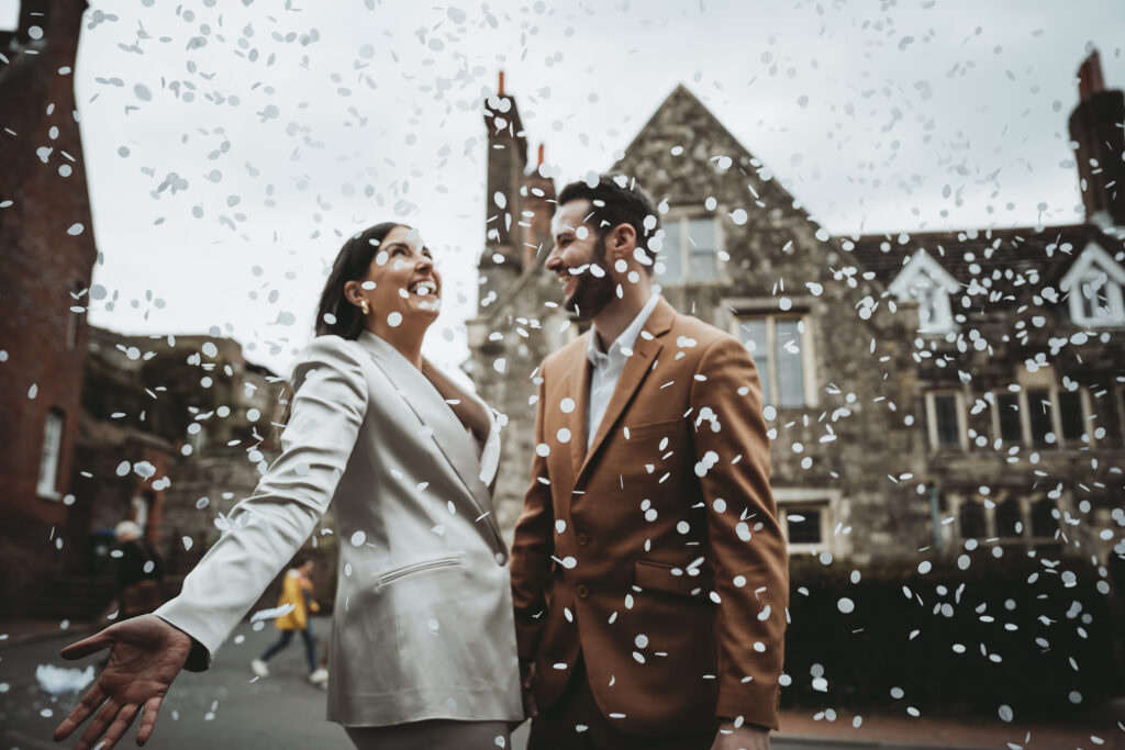 Laughing couple stand in front of a building as white confetti falls all around them.