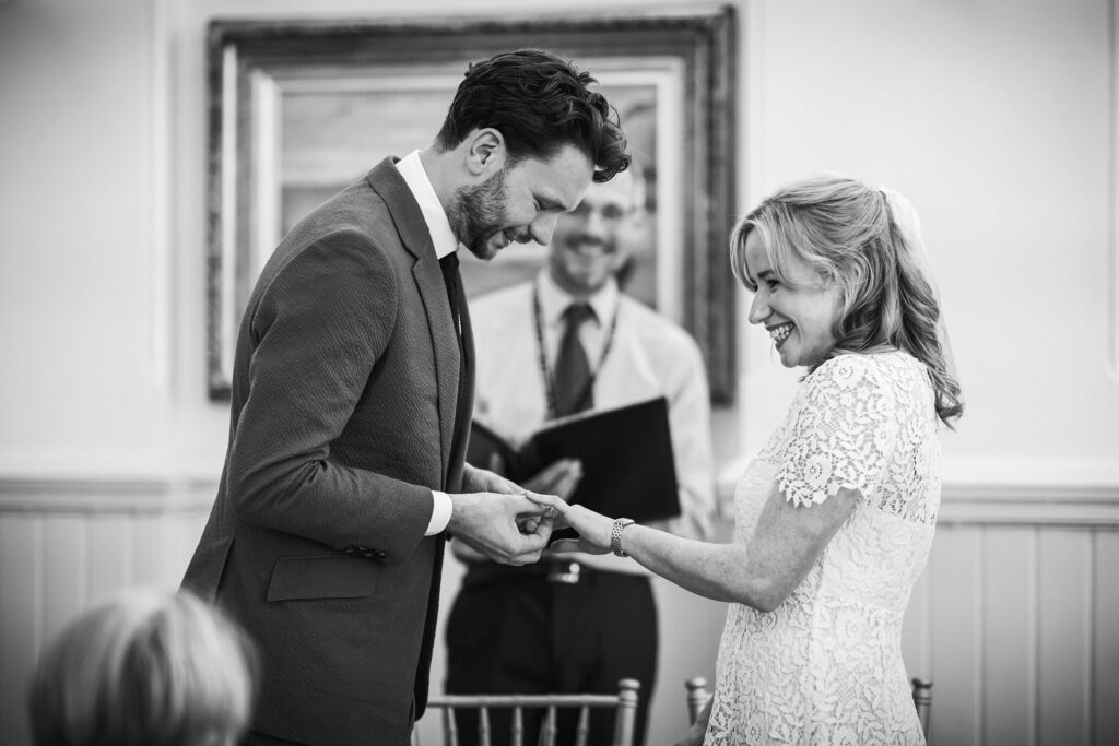 Black and white photo of the groom placing the wedding ring on the bride's finger during the ceremony.