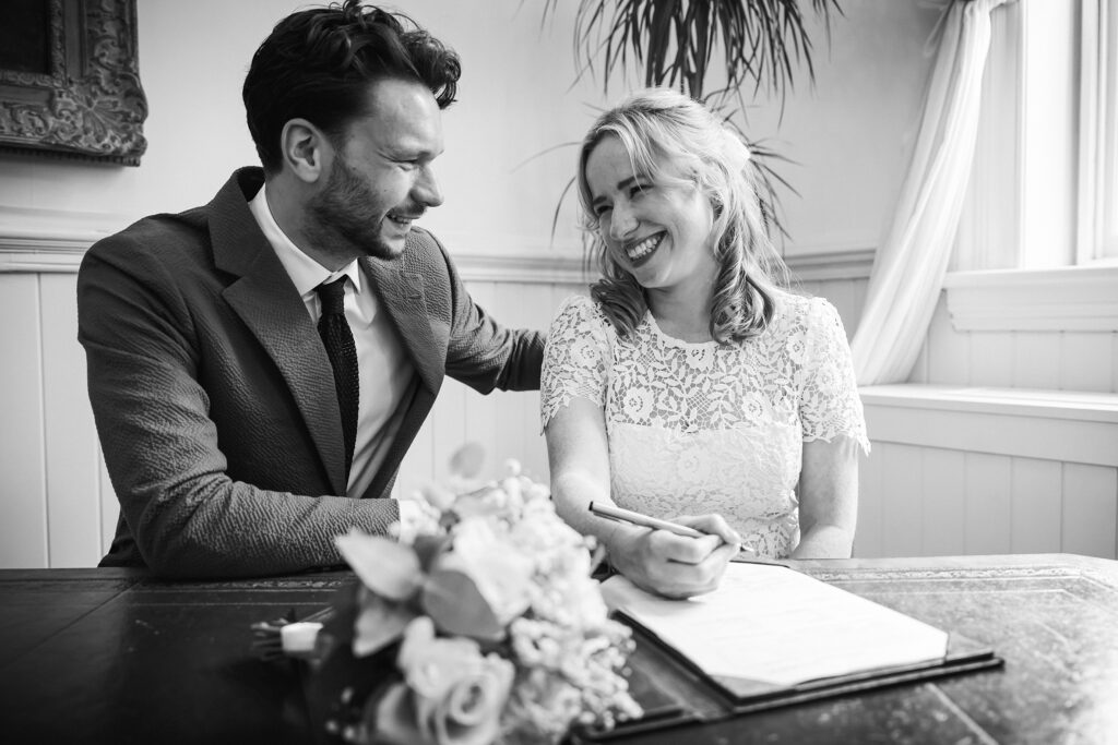 Black and white photo of the wedding couple signing their marriage certificate while smiling at each other.