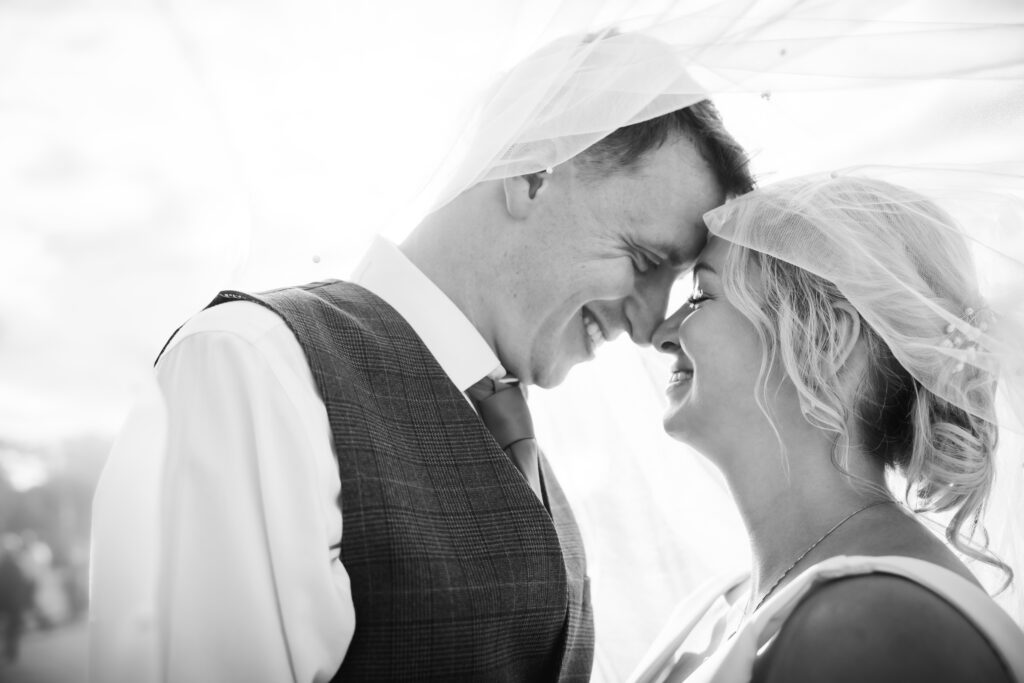 Black and white photo of the wedding couple smiling at each other beneath the bride's veil.