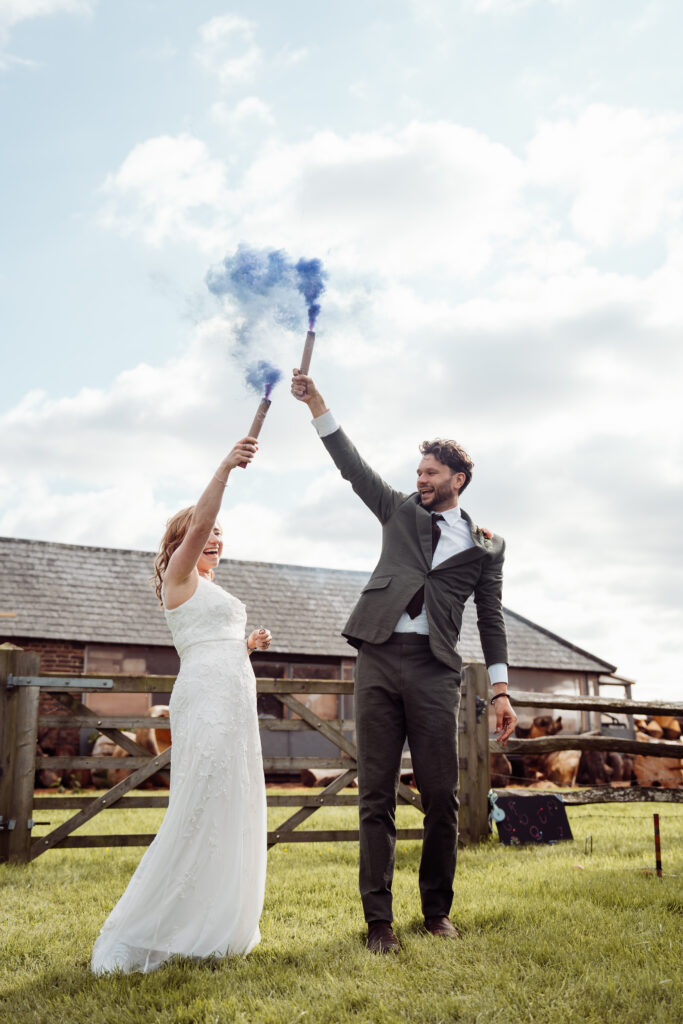 Bride and groom holding blue smoke flares above their heads outside.