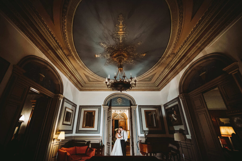 Bride and Groom embracing in the doorway of an ornate historical looking room with an imposing chandelier.