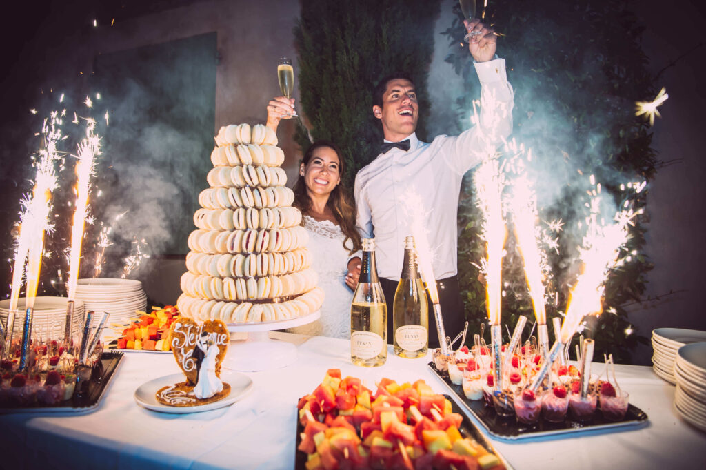 Smiling Bride and Groom, stood in front of a plate piled high with macaroons, raise their glasses.