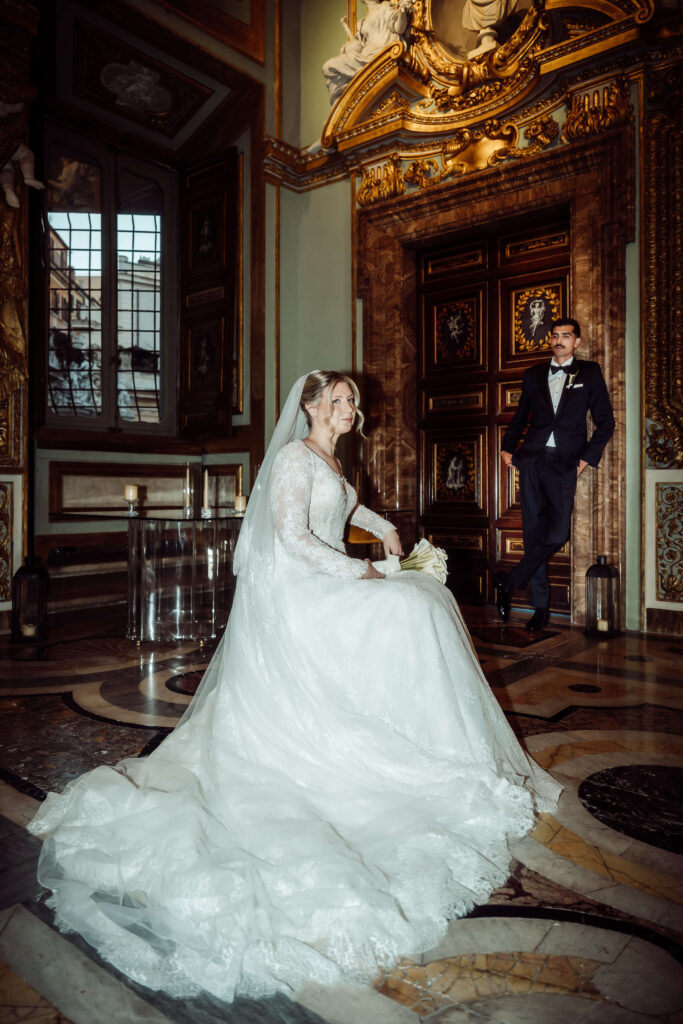 Bride seated in an ornate room with the groom leaning against the wall in the background.