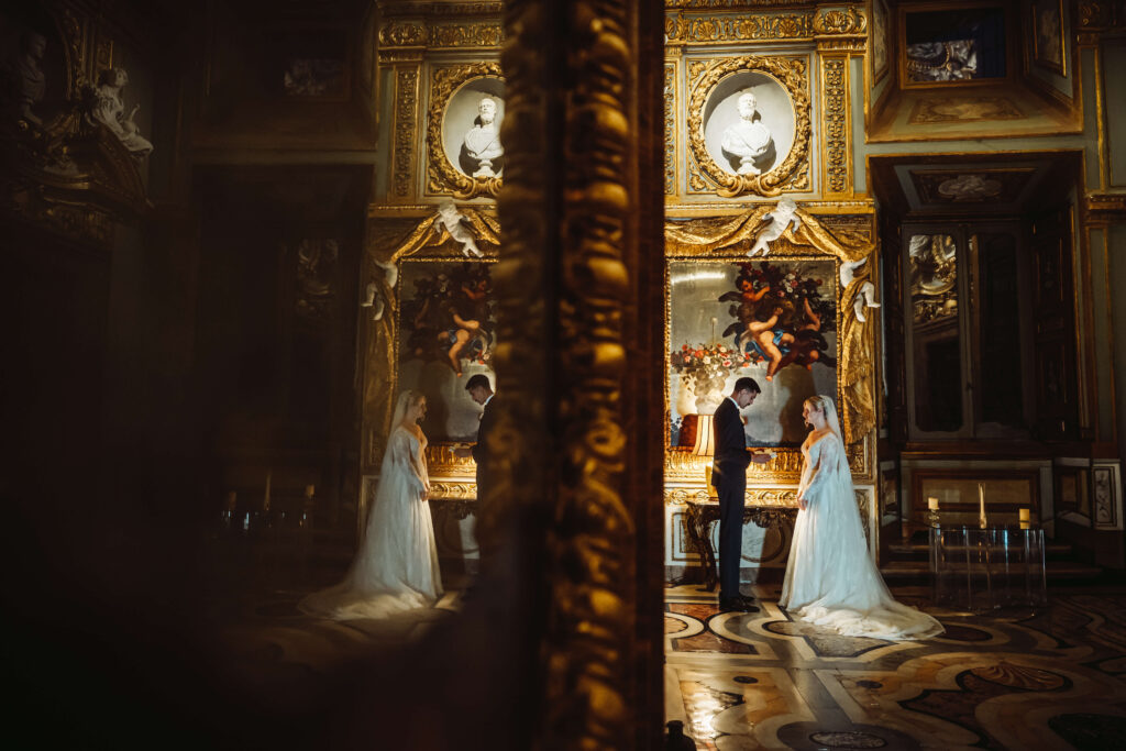 Groom reading from a piece of paper to the bride in an ornate room with gold architectural details.