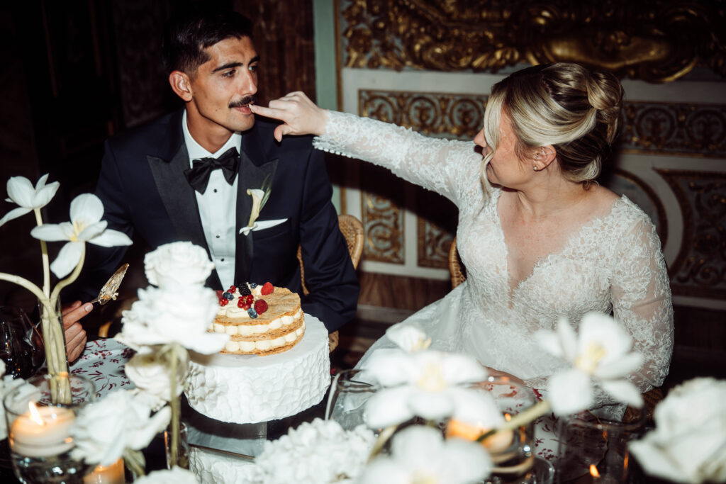 Sat in front of a cream layered cake, the Bride touches her Grooms lips.