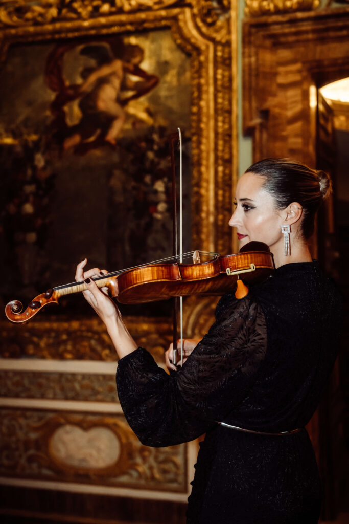 A violinist in a black dress plays in front of a Renaissance-style painting in an ornate golden frame.