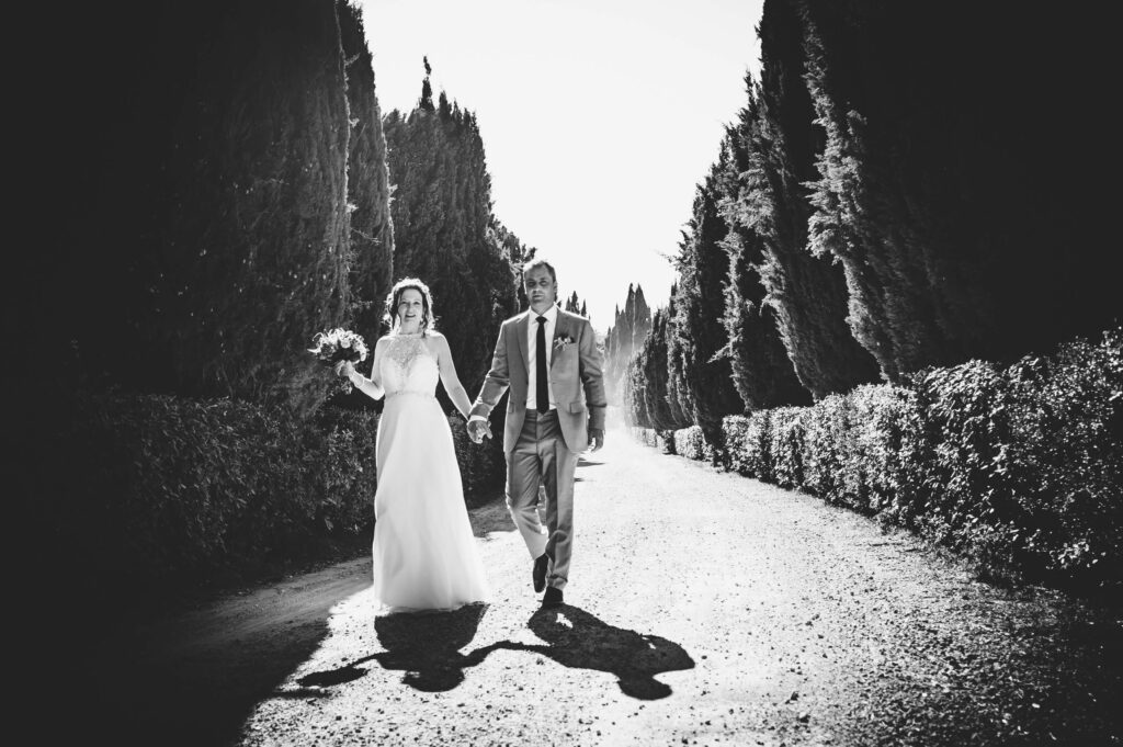 Black and white photograph of a Bride and Groom walking hand in hand along a treelined pathway.