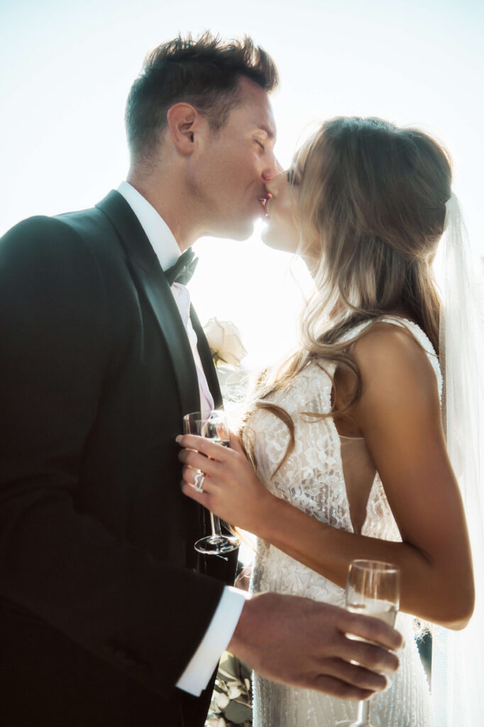 Wedding couple kissing while holding wine glasses with the sun behind them.