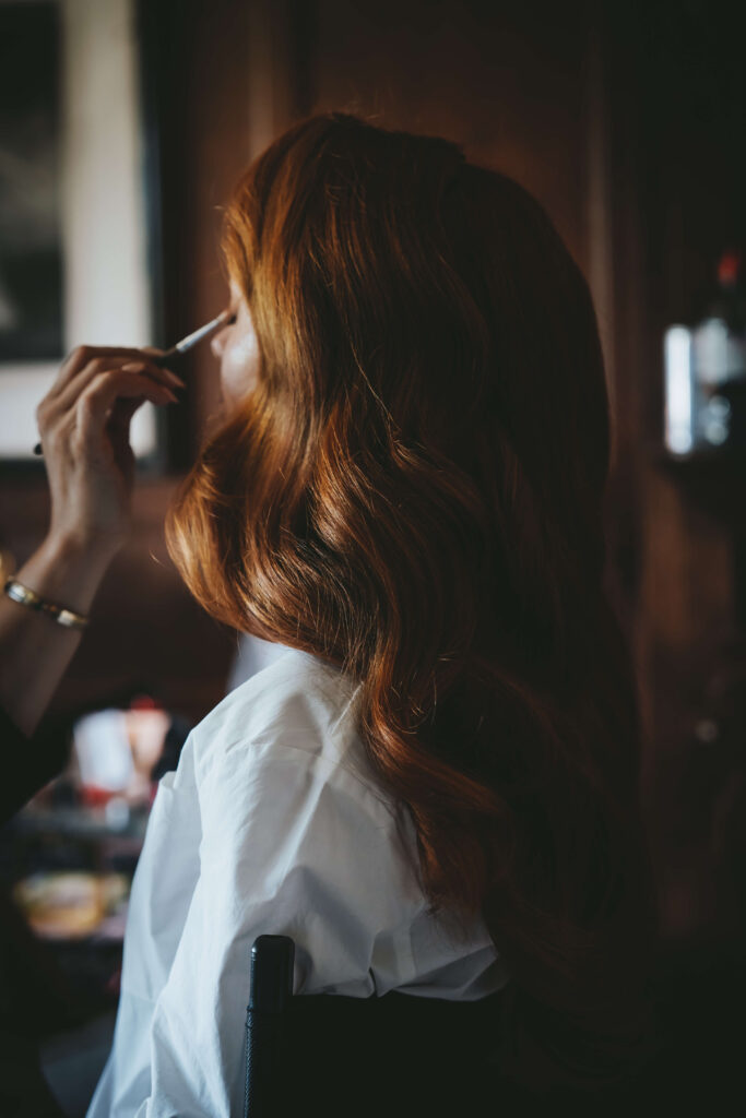Back view of a bride with curly ginger hair having her eye shadow applied.