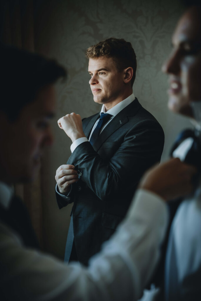 Groom adjusting his cufflinks while groomsmen help each other with their ties in the foreground.