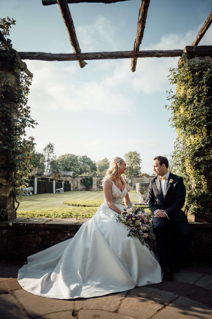 Wedding couple chatting while sitting on a low wall in front of a garden.