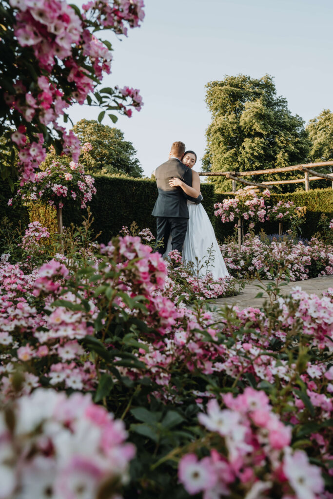 Bride and groom embracing in a floral garden with pink and white flowers.
