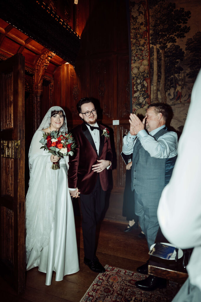 Wedding couple walking into a room, with the bride holding flowers and guests applauding in the corner.