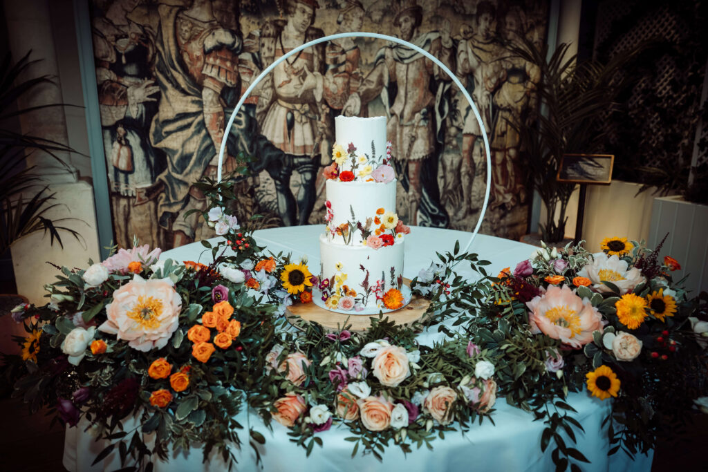 Three-tier wedding cake decorated with flowers on a table with a tapestry-style backdrop.
