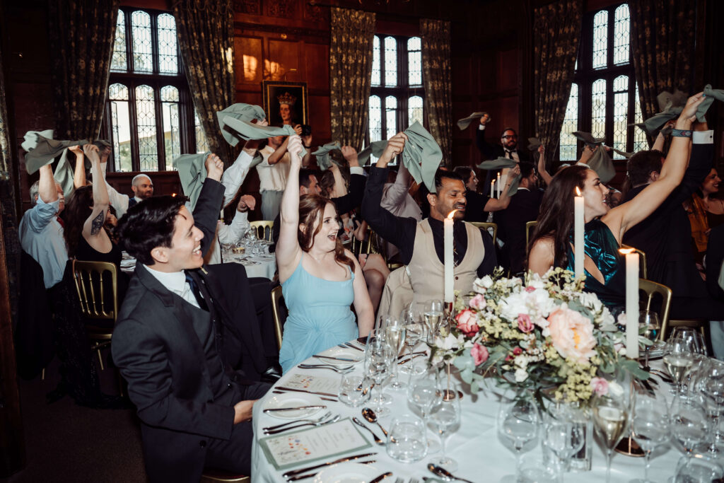 Wedding guests seated around decorated round tables at a reception, cheering and holding up their napkins.