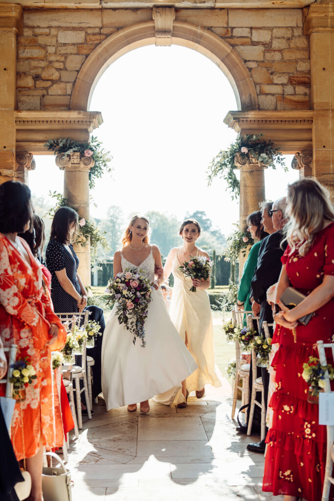 Bride and bridesmaid walking arm in arm down the aisle as wedding guests watch.