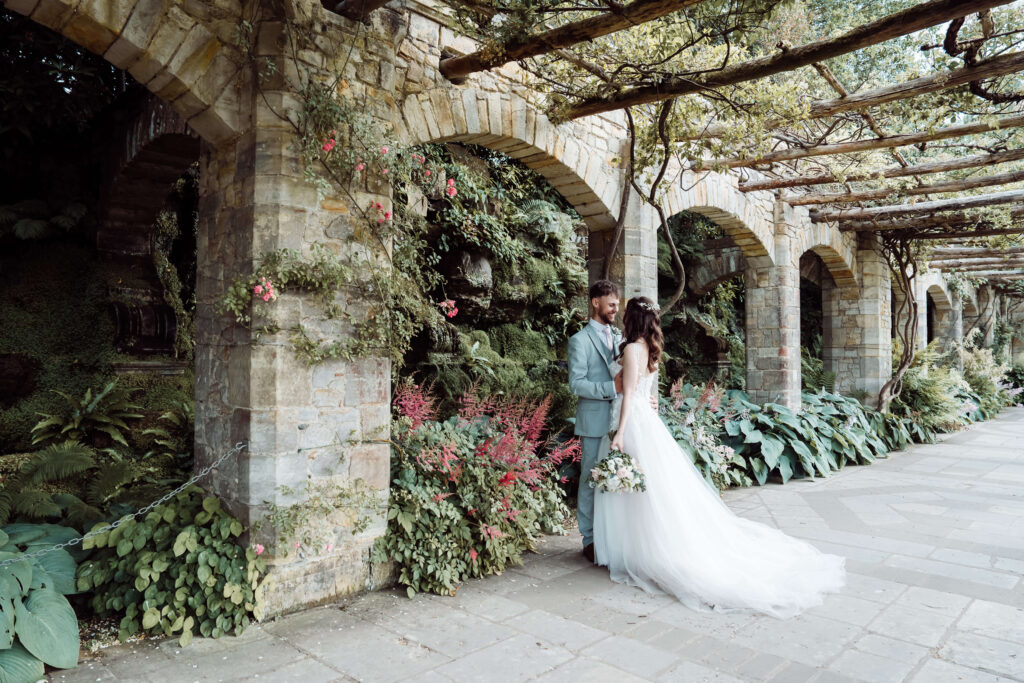 Bride and groom sharing an embrace next to stone arches and trailing vines.