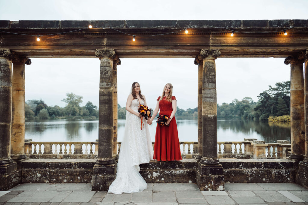 Two women standing under a stone colonnade by a lake holding bouquets, dressed in formal wedding attire.