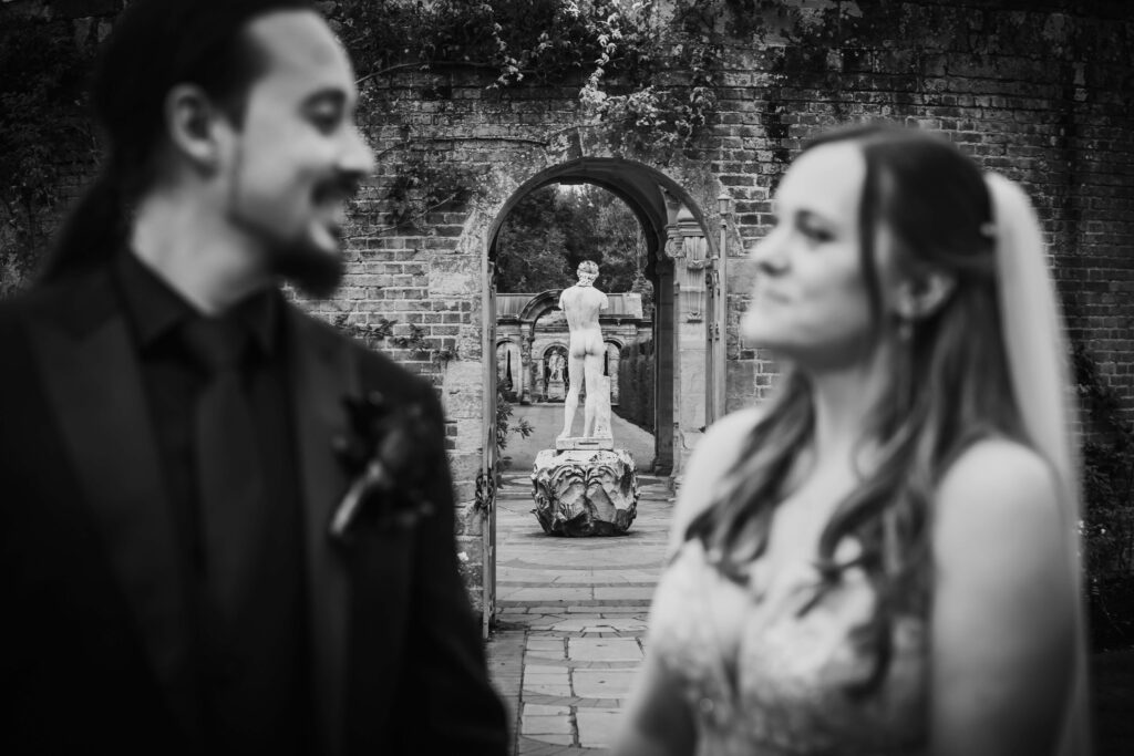 Wedding couple out of focus in the foreground looking at each other, with the arches and statues of Hever Castle gardens in sharp focus behind them.