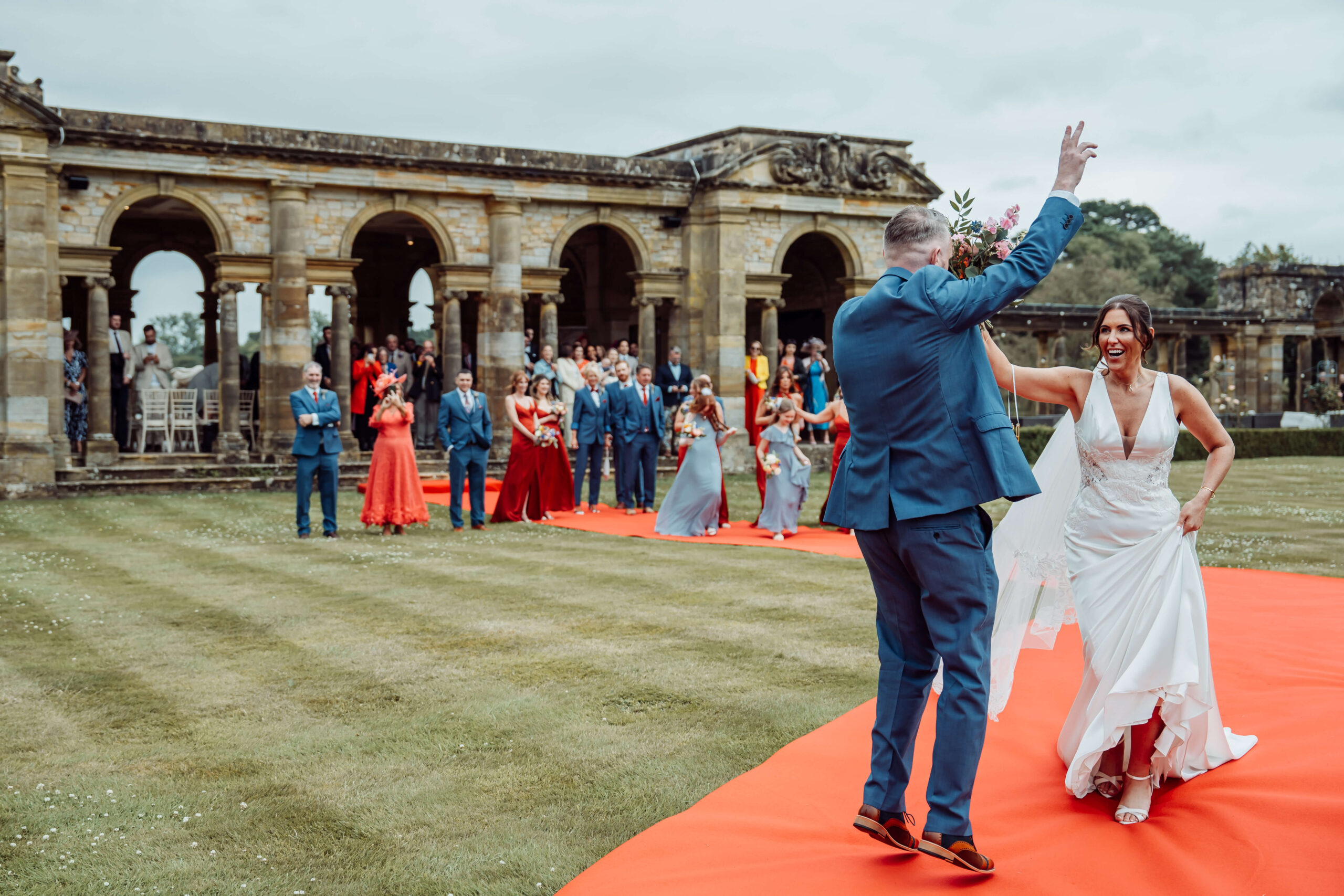 A wedding couple dances down the Hever Castle gardens along a red walkway, with guests watching from behind.