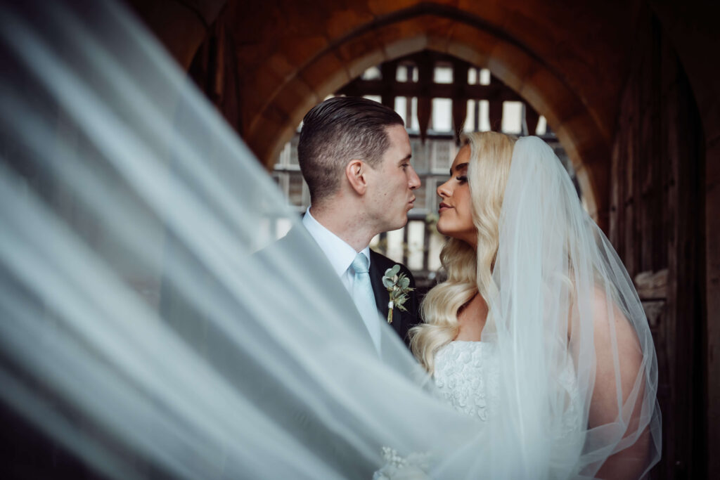 Bride and groom standing in front of a portcullis, looking at each other, with the bride's veil flowing across the foreground.