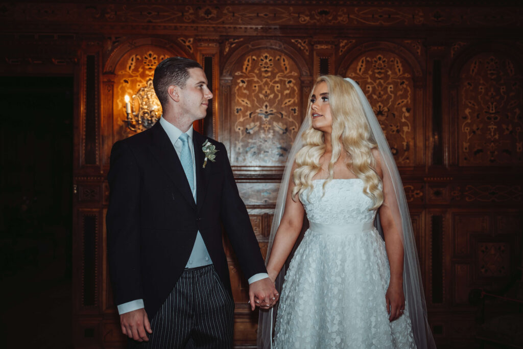 A Bride and Groom stand hand in hand, looking at one another, in front of an ornate wooden wall.