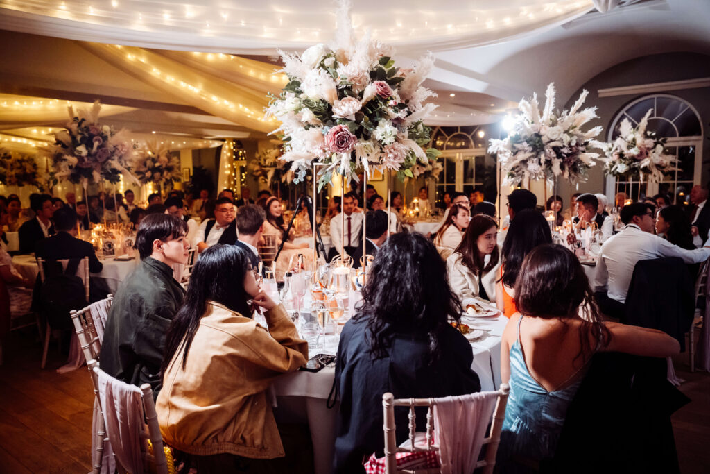 Wedding guests seated at decorated round tables with flowers, food, and glassware.