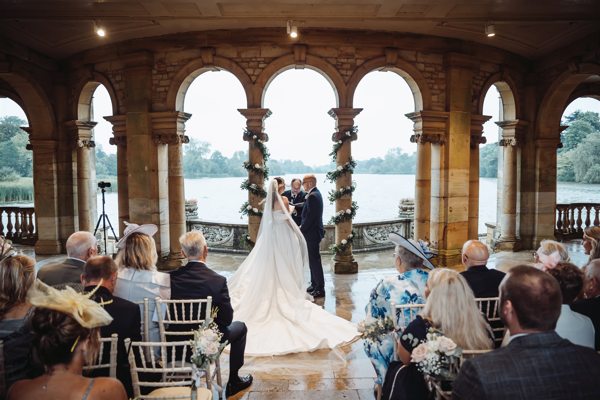 Wedding couple during their ceremony at Hever Castle Loggia, with the lake behind them and guests seated in front. [Teri V Photography Wedding Timeline Guide]