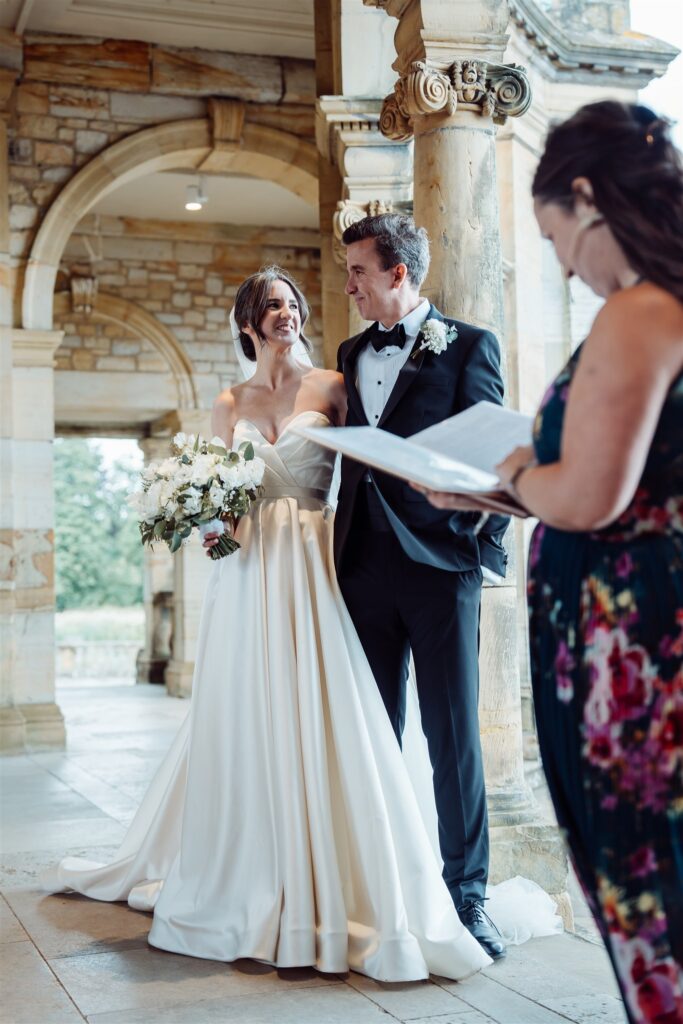 Wedding couple standing during their ceremony while an officiant reads from a script in an ornate stone courtyard.