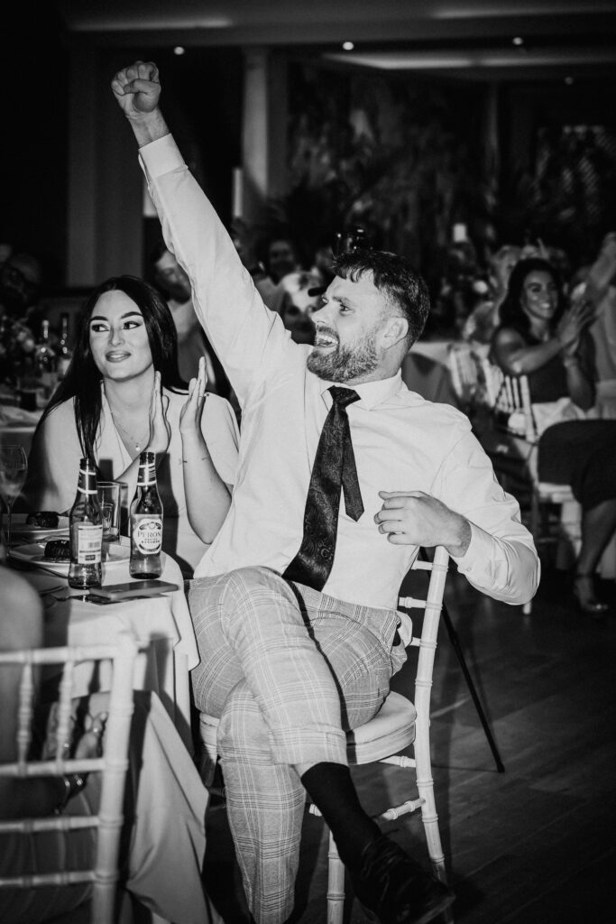 Black and white photo of a wedding guest cheering and raising his arm during the reception while seated at a table.