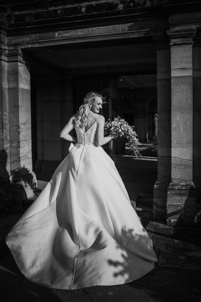 Black and white photo of the bride from behind as she walks through a stone archway, smiling over her shoulder.
