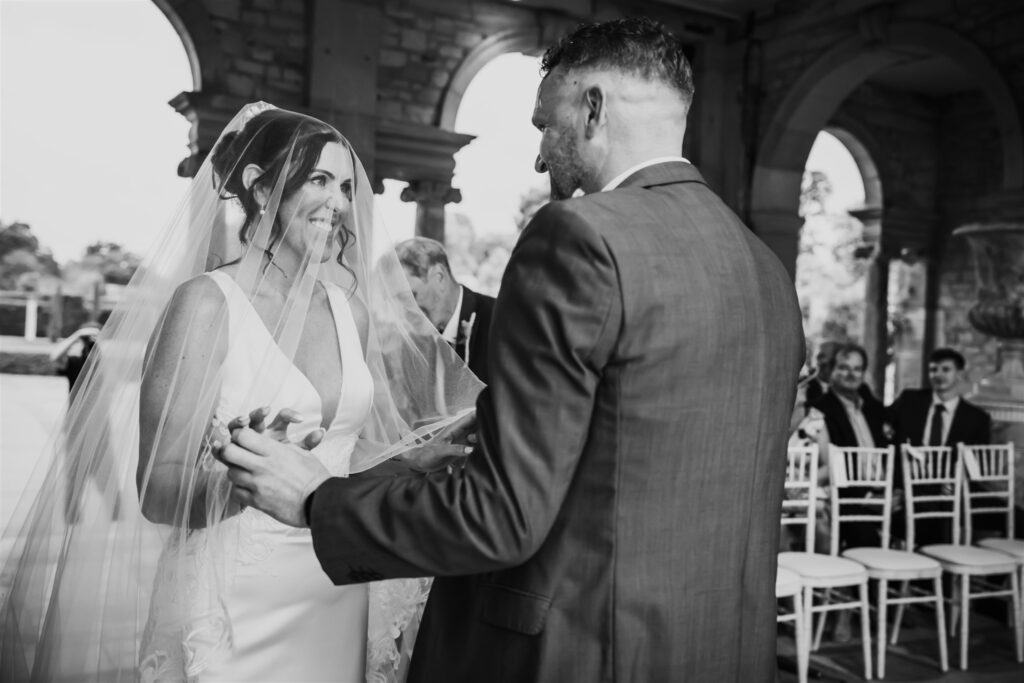 Black and white photo of the wedding couple smiling at each other during their ceremony.