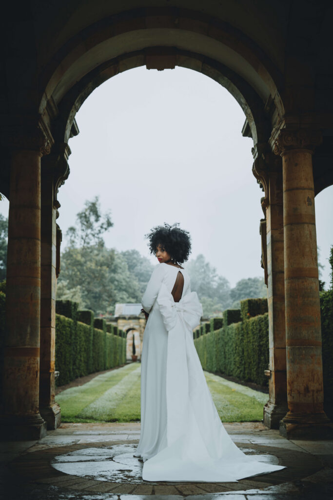 Bride looking over her shoulder at the camera while standing beneath a stone archway, with a garden behind her and a large white bow on the back of her dress.