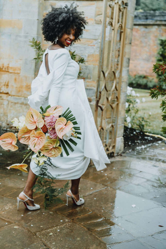 Bride in a white dress holding a bouquet of flowers, smiling while walking past a stone building.
