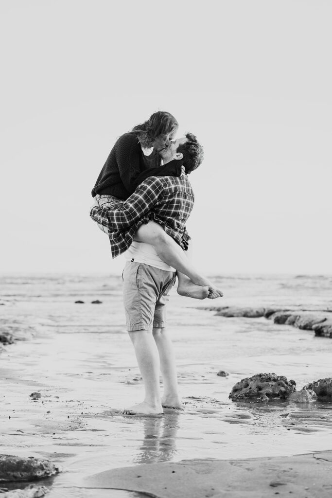 Black and white photo of the groom holding his fiancée in his arms as they kiss, facing each other.