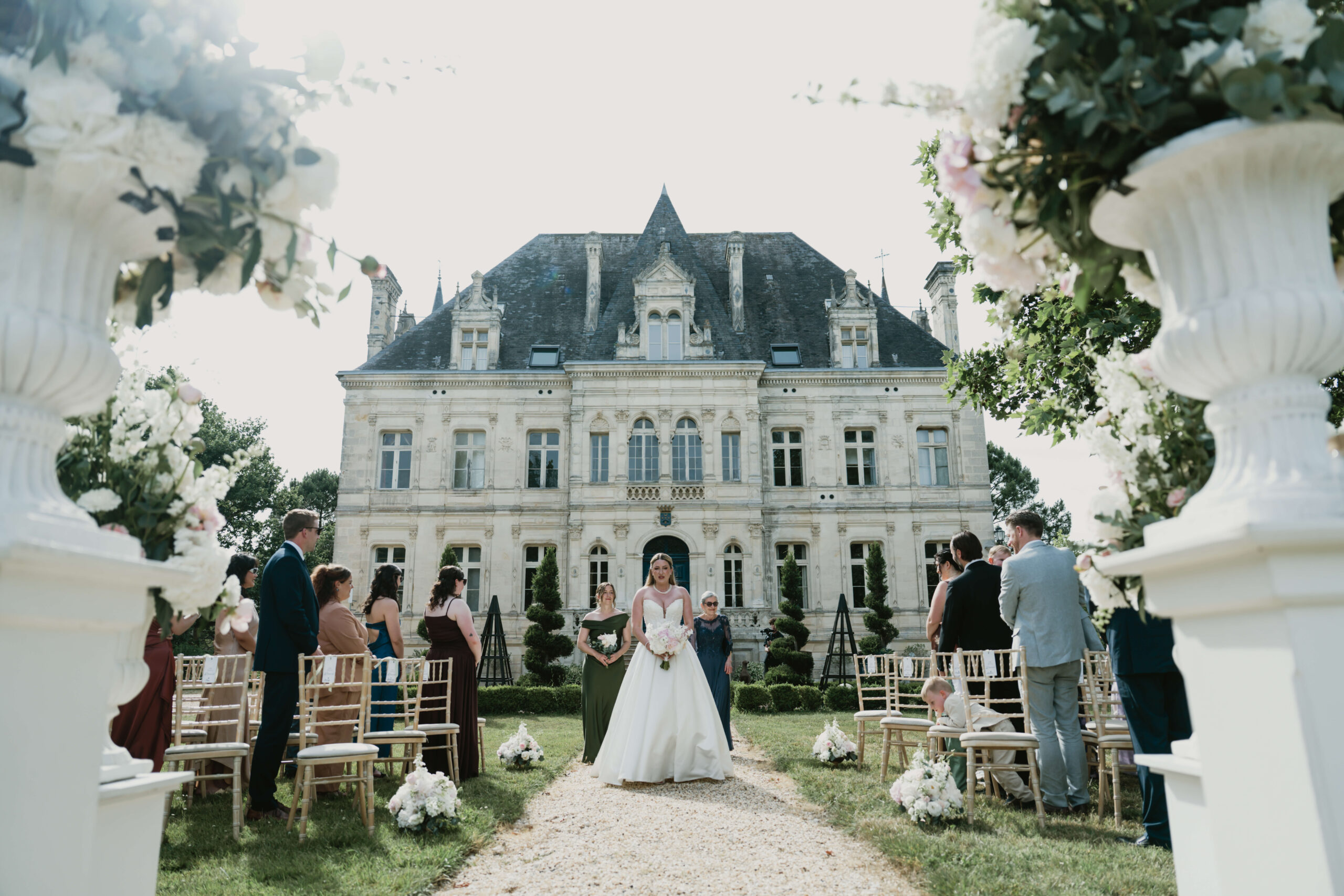 Bride wearing a white dress and walking down the isle to her wedding ceremony in front of Chateau De La Valouze in France.