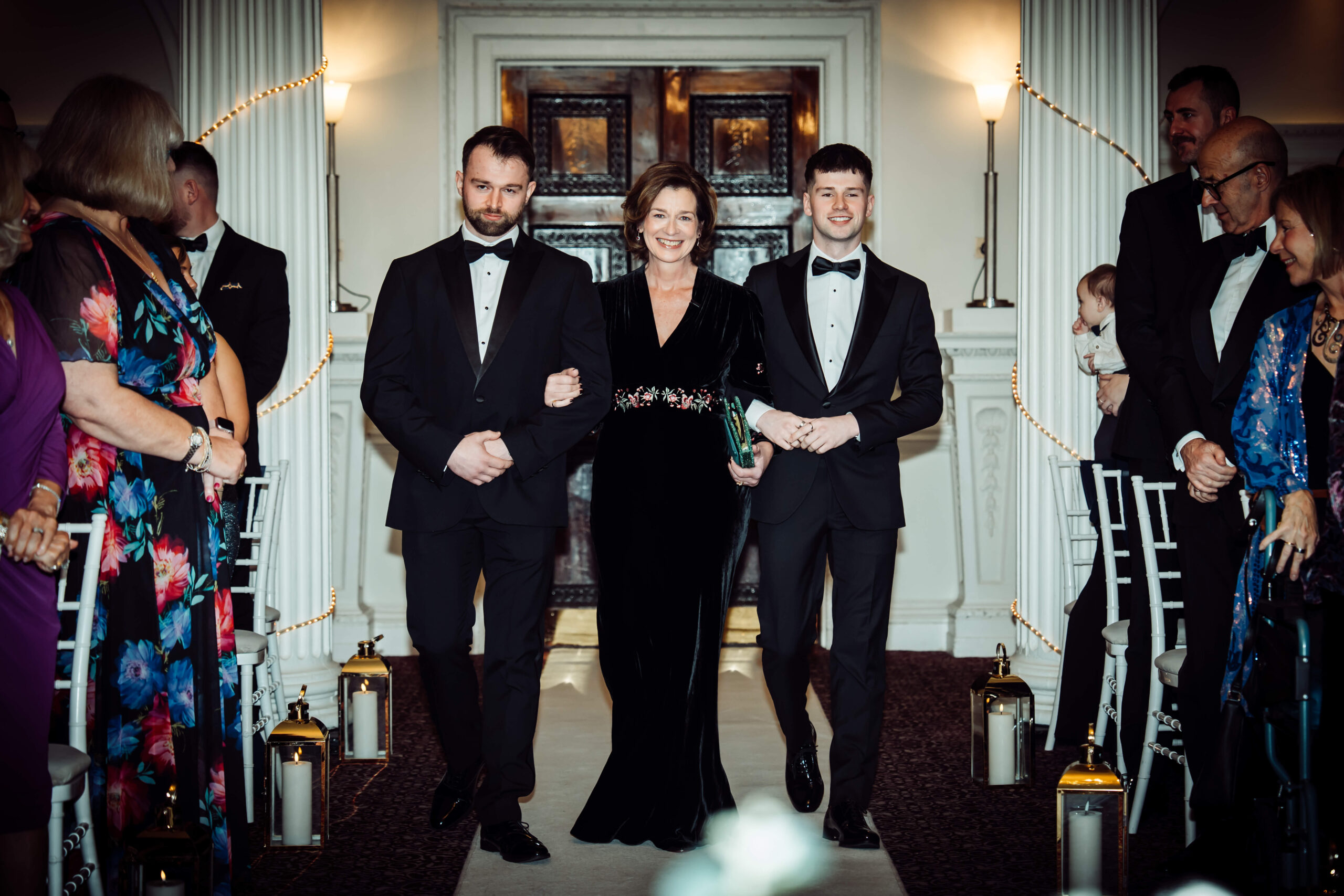 Two men and one woman are walking down the aisle, with guests standing from their seats.