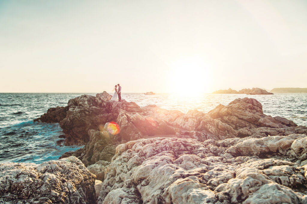 a couple stands on a rock jutting out to see on their wedding day. The Croatian sunset is in the background.