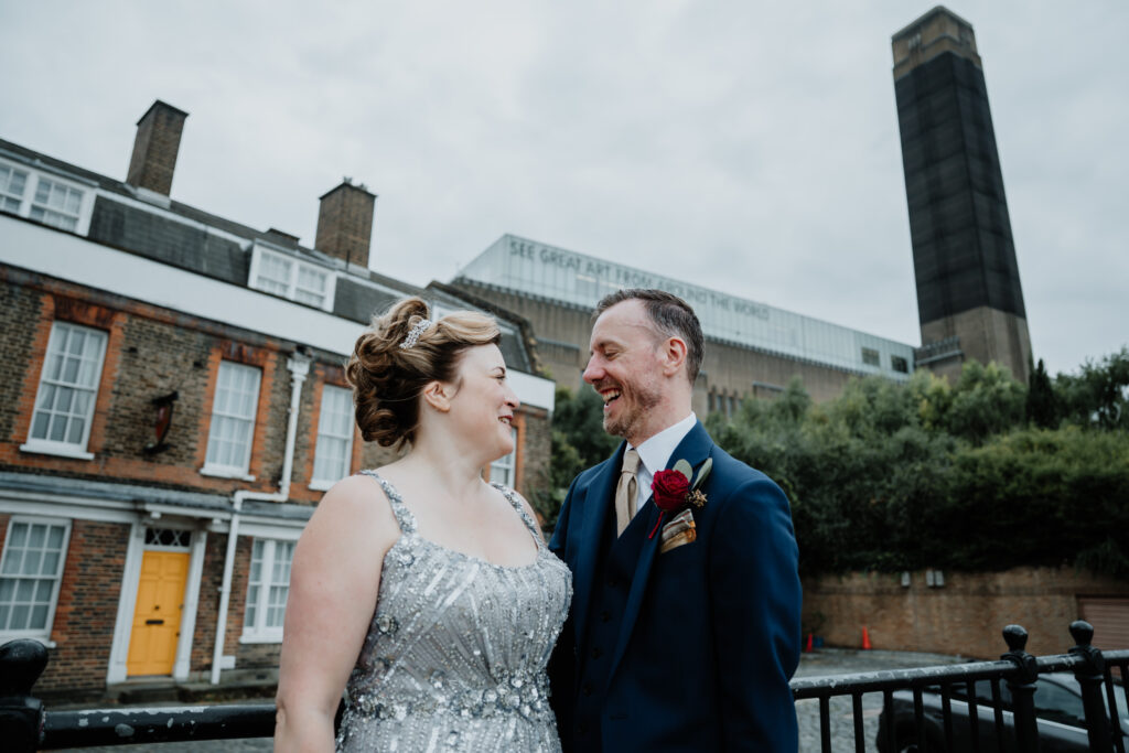 Couple smiling and laughing towards each other in a wide shot. Captured by a UK luxury wedding photographer in front of the Tate Modern.