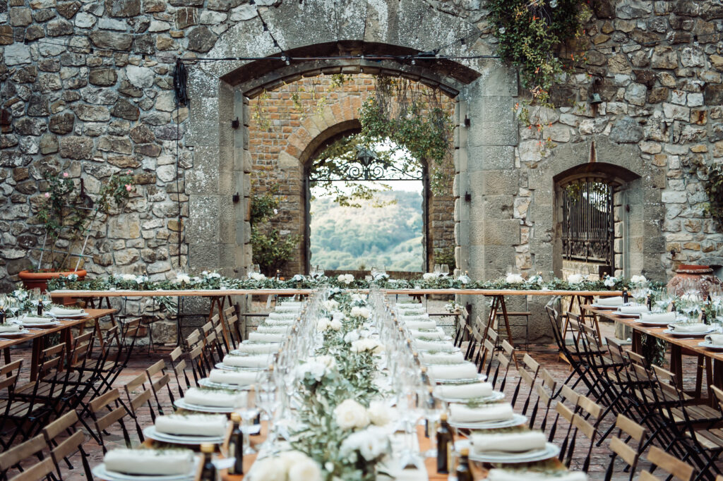 Elegant wedding reception tables decorated with white flowers and classic tableware in the stone courtyard of Castello di Cafaggio, Italy