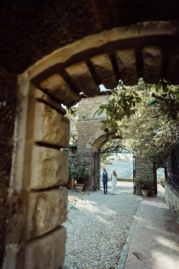 Bride and groom walking through castle grounds, framed by a stone archway.