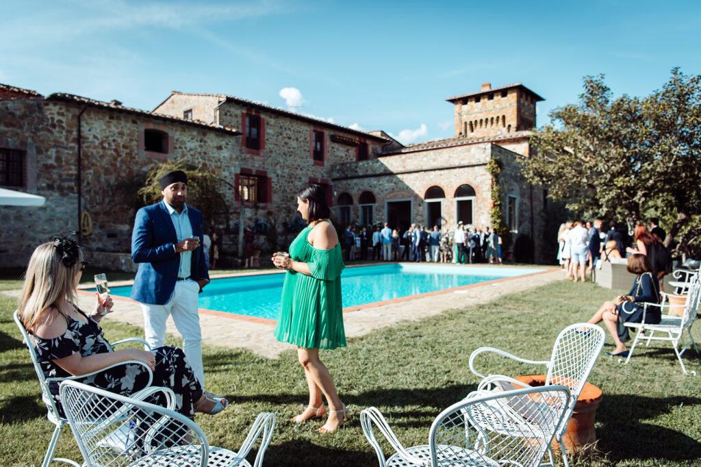 Wide shot of guests standing, chatting, and enjoying drinks by the pool outside of Castello di Cafaggio