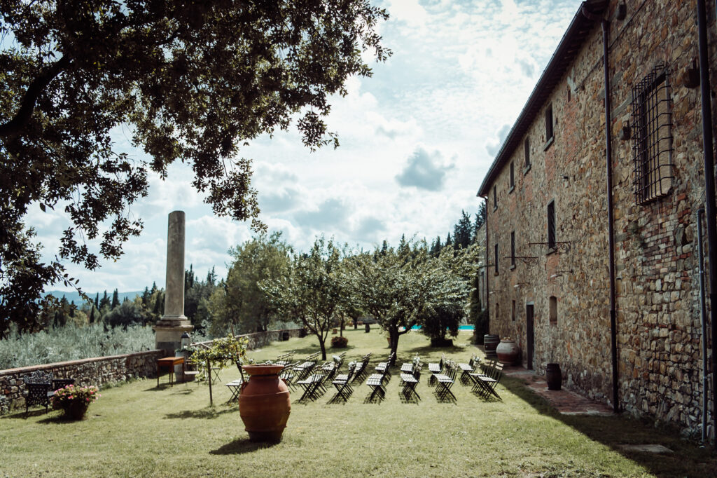 Outdoor wedding ceremony setup at Castello di Cafaggio, with chairs and floral arrangements, the castle side visible, and panoramic Tuscan hills and trees in the background