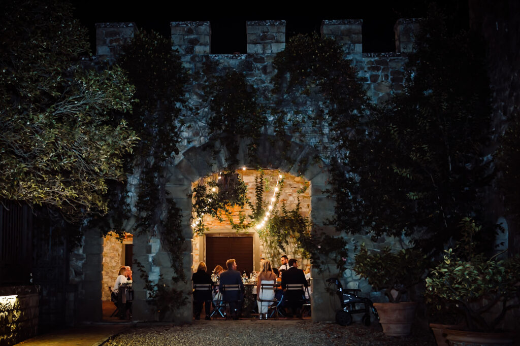 Wide shot of Castello di Cafaggio with a stone archway framing guests at their tables, lit by fairy lights during the Castello di Cafaggio wedding