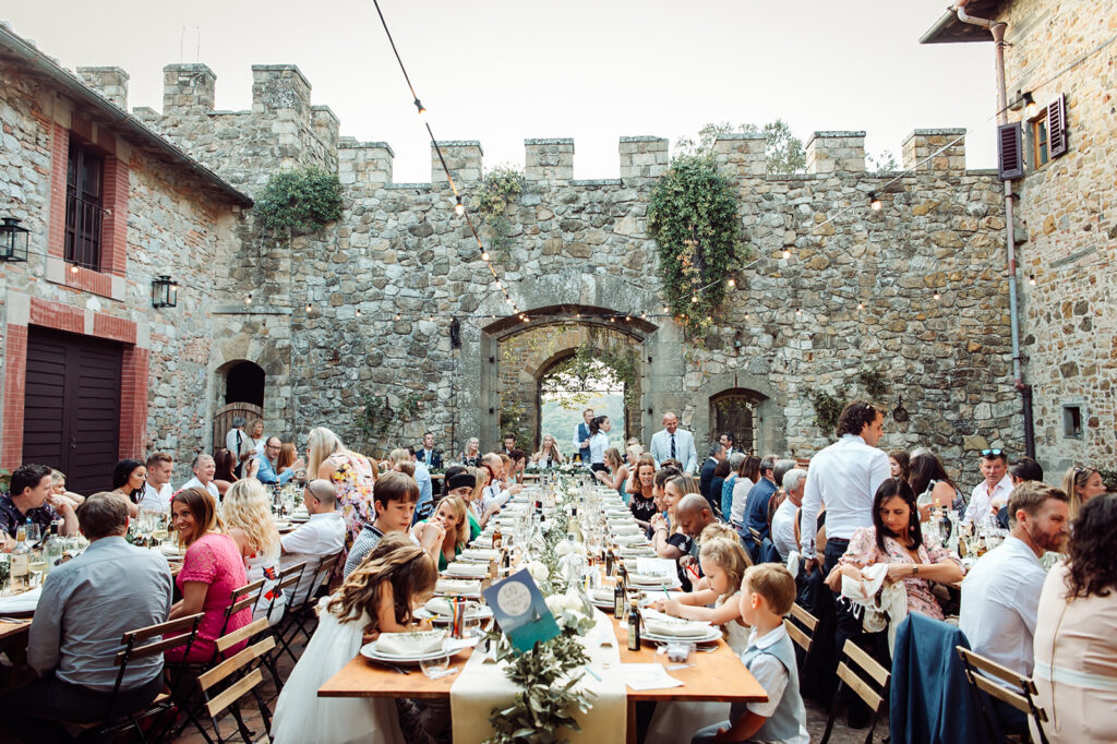 Wide shot of wedding guests seated and chatting at long tables in the castle courtyard during the Tuscan wedding